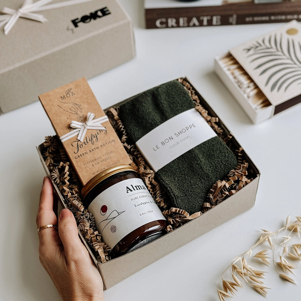 The Perdy Gift Box byFoke laying open on a table with a woman's hand holding the box. The gift box contains; Cloud Socks by Le Bon Shoppe, Alma Essential Oil Soy Wax Candle, Fortifying Green Bath Potion by MOA