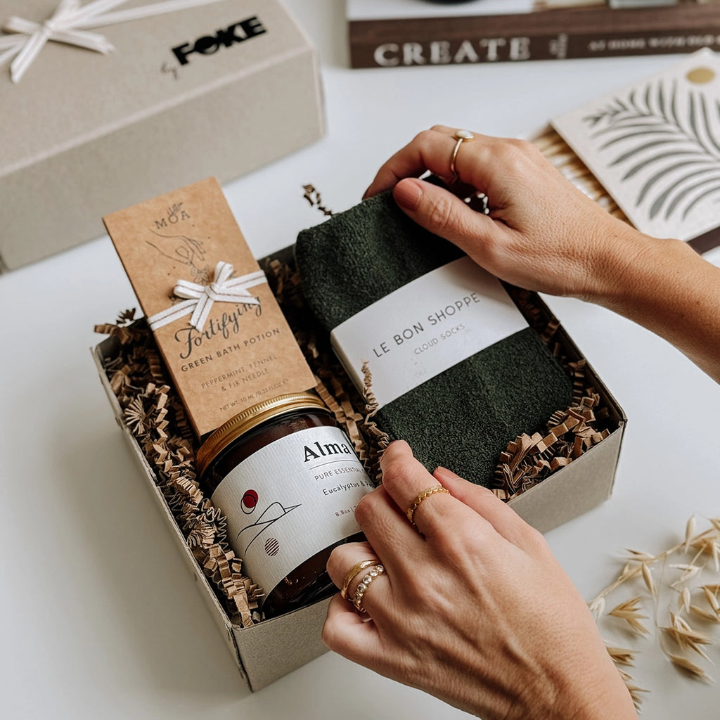 The Perdy Gift Box byFoke laying open on a table, a woman's hands are placing the socks in the box. The gift box contains; Cloud Socks by Le Bon Shoppe in Forest, Alma Essential Oil Soy Wax Candle, Fortifying Green Bath Potion by MOA