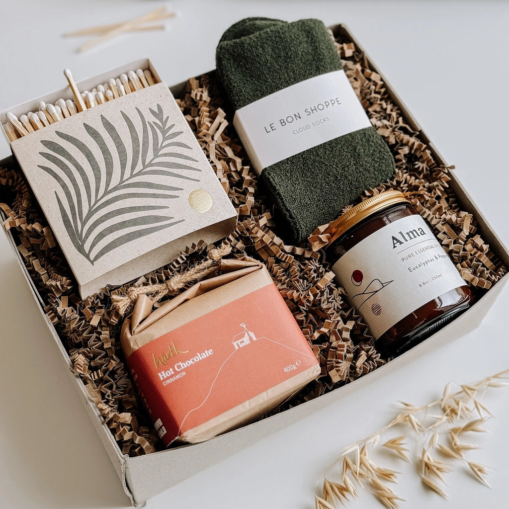 A table displays a gift box featuring Le Bon Shoppe socks, an Essential Oil Soy Wax Candle, a box of luxury matches in a letterpress printed box, and Hearth Hot chocolate.