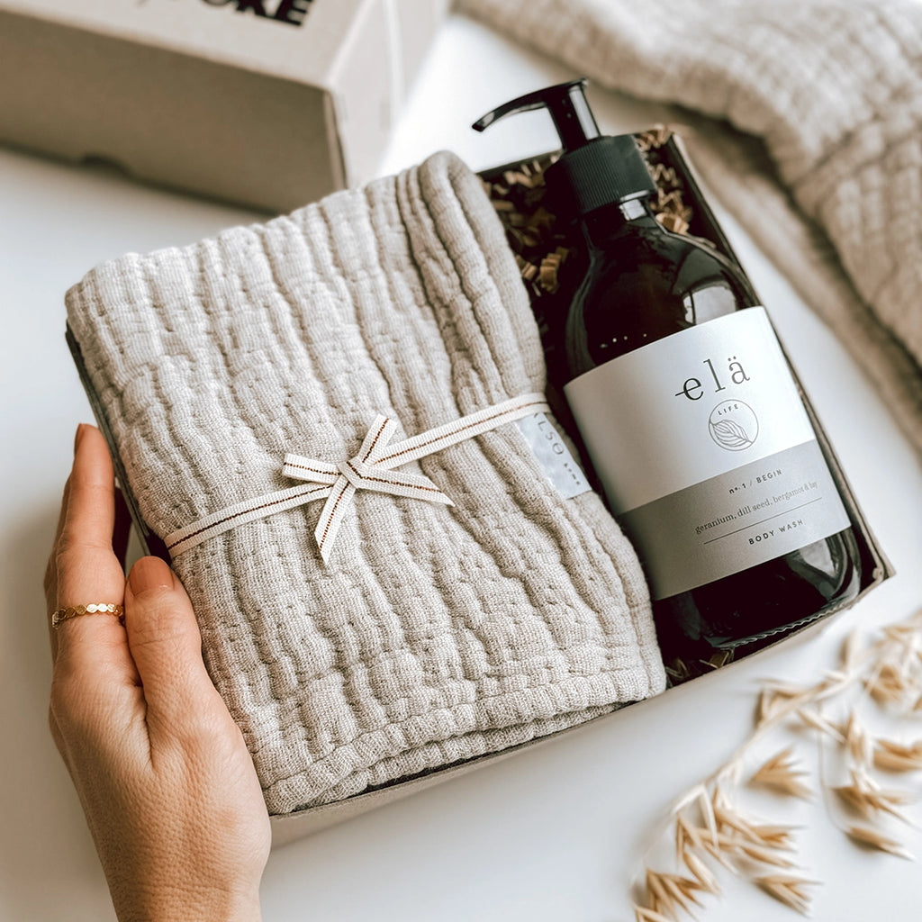 A woman has opened the Una Gift Box byFoke, she is touching the box with her left hand. The box is lying open with the lid to one side. Inside the box you can see Ela Life Body Wash and a Textured Wash Cloth.
