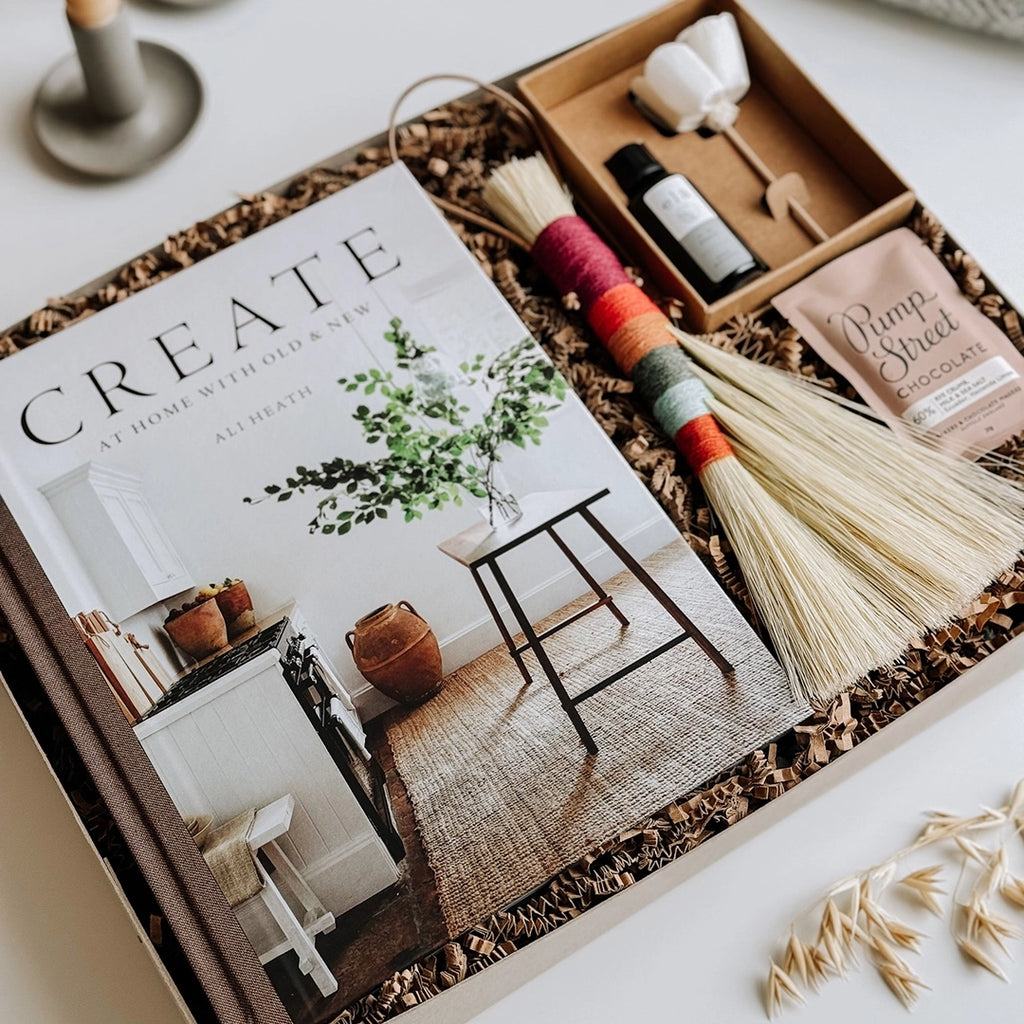 The Isla Gift Box byFoke is open on a table displaying its contents; the book Create by Ali Heath, a Twig Stick Tampico Brush, an Essential Oil Flower Diffuser and a bar of Pump Street Chocolate.