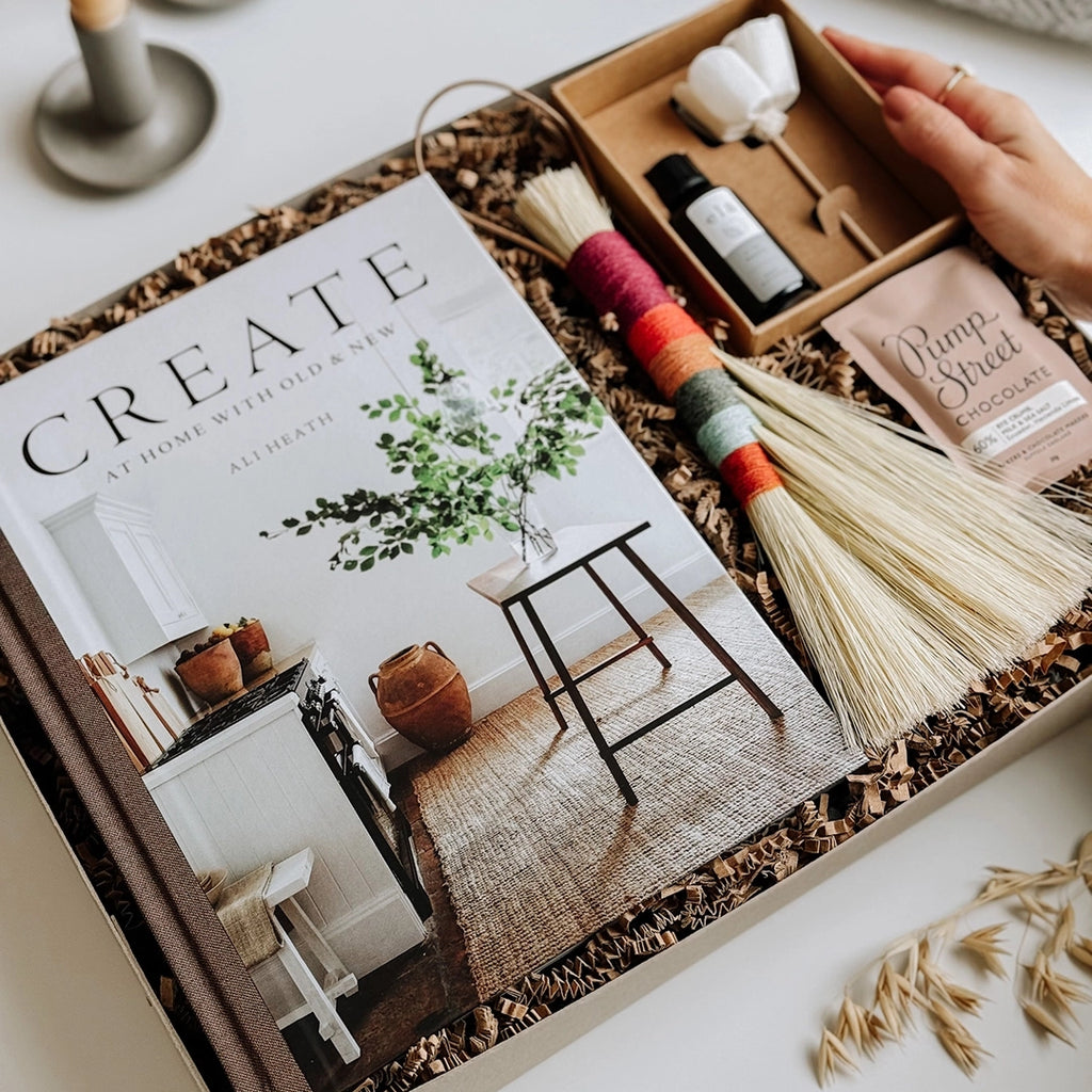 A woman's hand is placing one of byFoke's Isla Gift Boxes on a table. Inside the box you can see the book Create by Ali Heath, a Twig Stick Tampico Brush, an Essential Oil Flower Diffuser and a bar of Pump Street Chocolate.