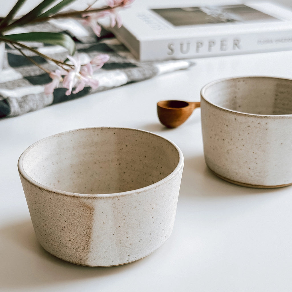 A close up of a pair of hand thrown stoneware bowls made by Clai. The bowls are on a table in the byFoke gift shop. they have an off white glaze with a brighter white stripe running through.