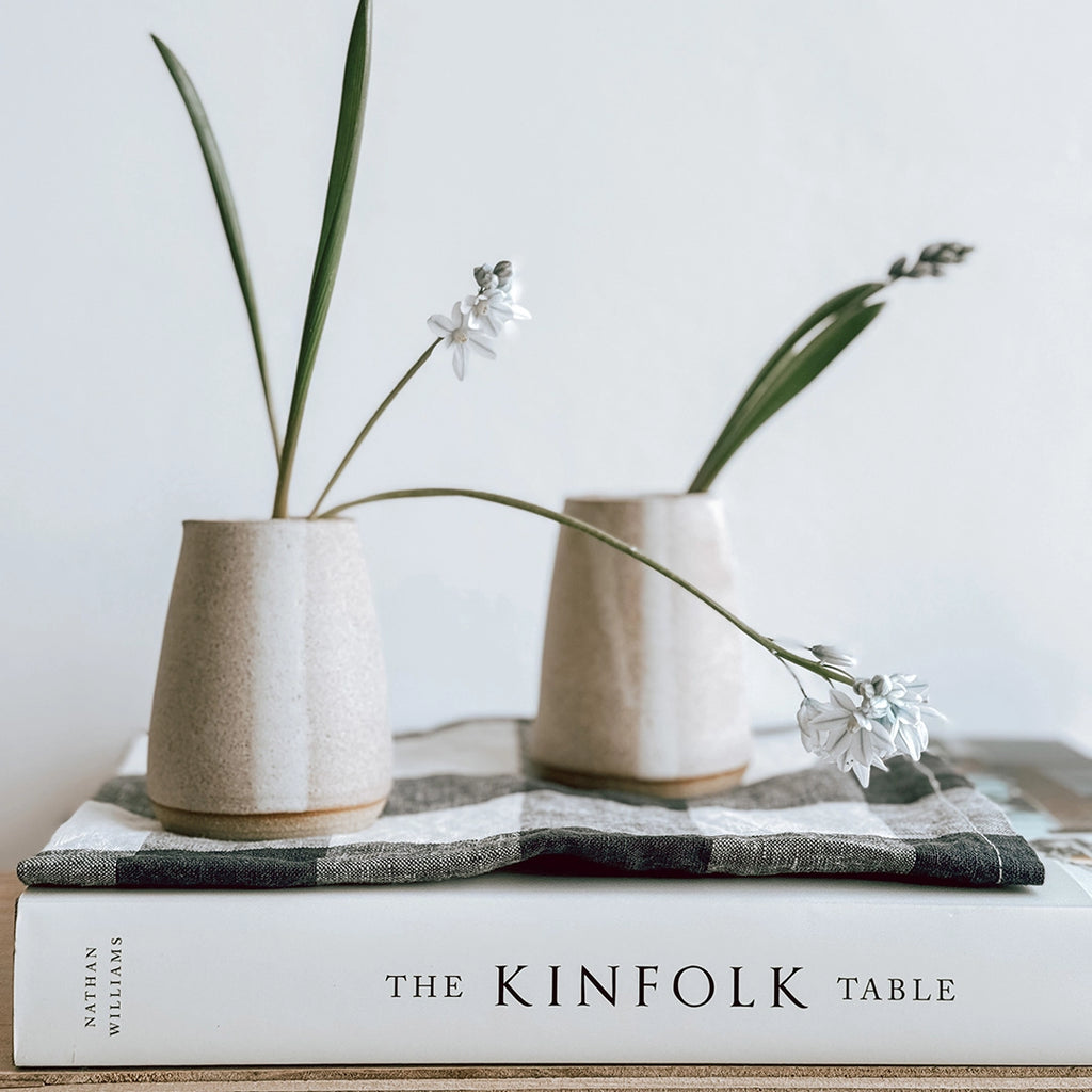 A pair of Hand thrown stoneware jugs with spring flowers inside dispayed ona shelf on top of a gingham napkin and The Kinfolk Table Recipe book.