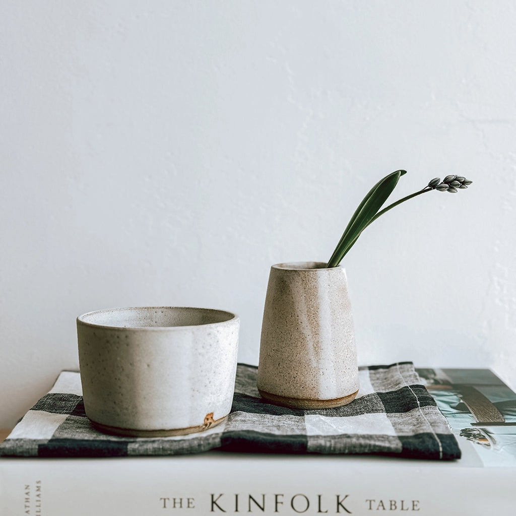 A hand thrown stoneware jug and matching bowl displayed on a shelf on top of a gingham napkin and The Kinfolk Table Recipe book. The jug has spring flowers inside. The photo was taken in the byFoke gifting studio.
