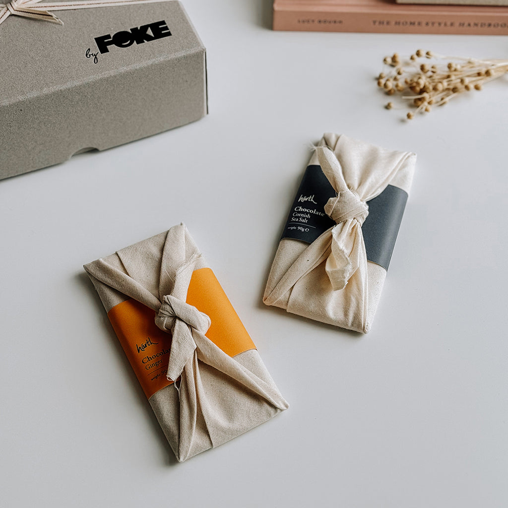 Two bars of Harth Chocolate, one ginger Chocolate, one Cornish Sea Salt Chocolate. The chocolate bars are wrapped in Harth's fabric Furoshiki wrapping.  The chocolate bars are laying on a table with a byFoke gift box and some dried flowers in the background.