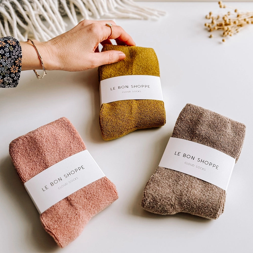 Three pairs of Cloud Socks by Le Bon Shoppe in 3 colours, Mulberry, Olive and Frappe. A woman's hand  is holding the corner of the olive pair of socks. byFoke.