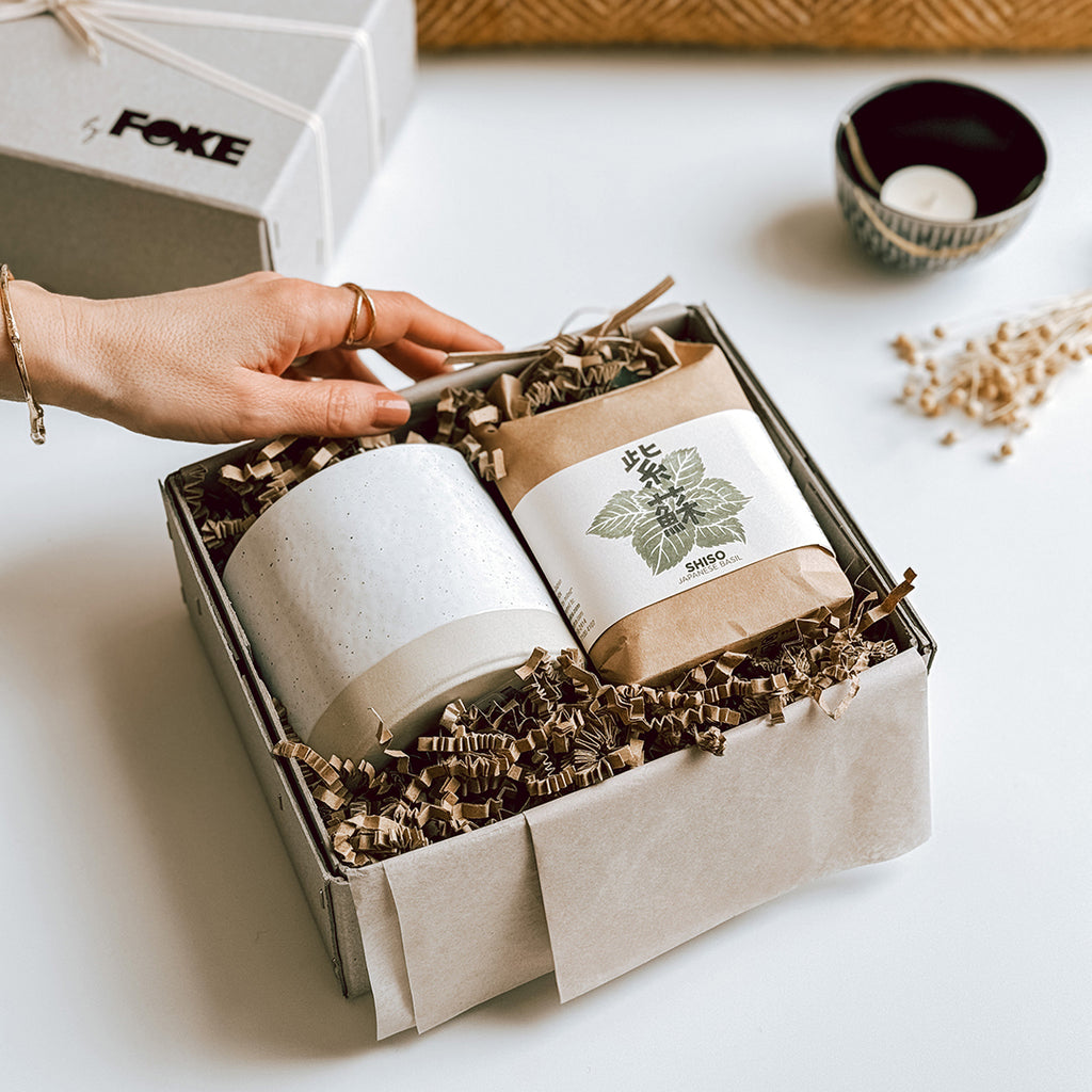 The Maiko gift box byFoke laying open on a table showing the contents, which are a Japanese Herb Growing Kit and a ceramic plant pot. A woman's hand is holding the edge of the gift box.
