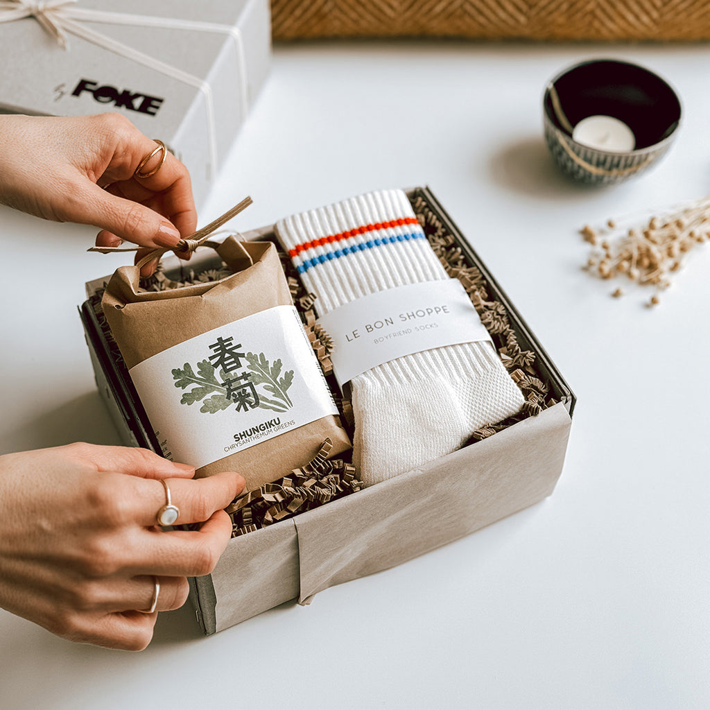 A woman's hands are placing one of the gift items into the byFoke Mei Gift box. The Gift box contains a pair of Le Bon Shoppe Boyfriend Socks and a cultivate and eat Japanese Herb Kit.