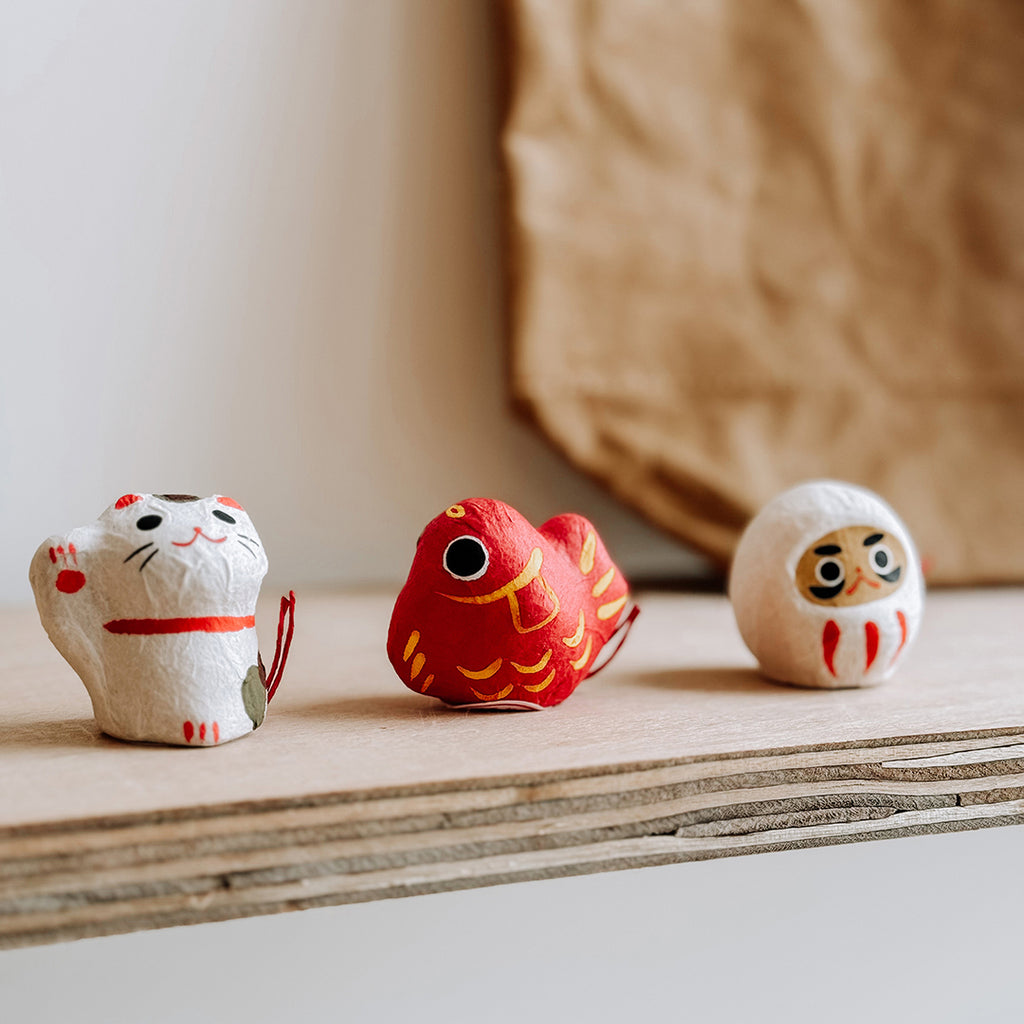 A close up of three Japanese Flowering Fortune Figurines on a shelf in the byFoke Gift shop. One is a Beckoning Cat, one is a Koi and one is a Daruma