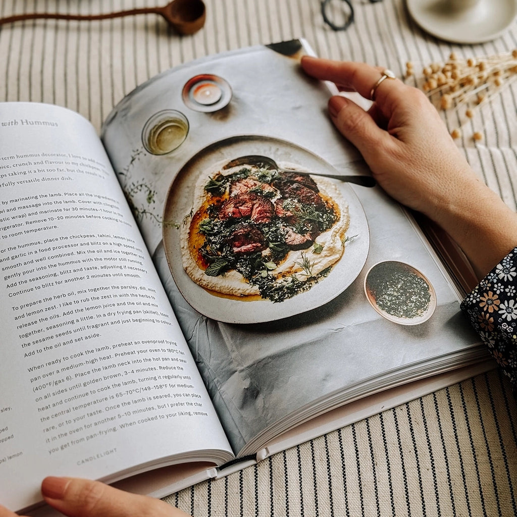 Supper, recipes worth staying in for by Flora Shedden. The cook book is being held open, showing one of the recipes inside. The book is on table with a striped table cloth. byFoke