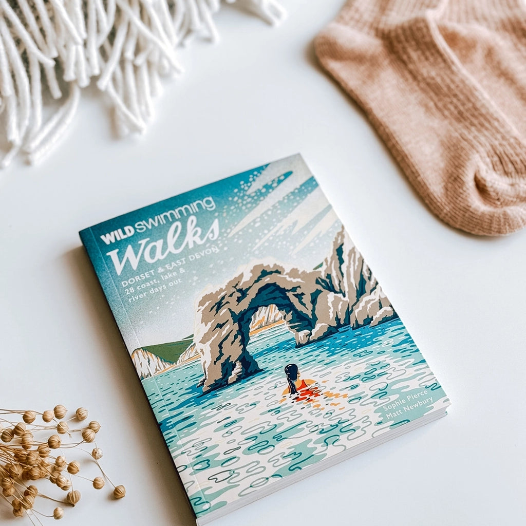 Wild Swimming Walks, Dorset & East Devon Guide Book, laying on a table next to a blanket and a pair of cottage socks. byFoke
