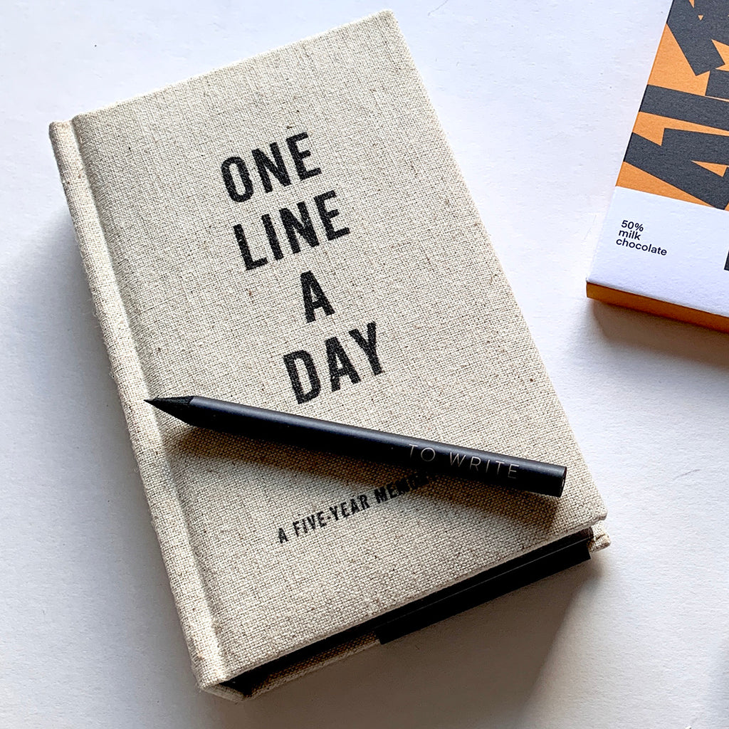 A photo of a One Line A Day Five Year Memory Book by Chronical books, laying on a table with a small black pencil laying on top of the journal, in the background you can see the corner of a bar of Ocelot chocolate.