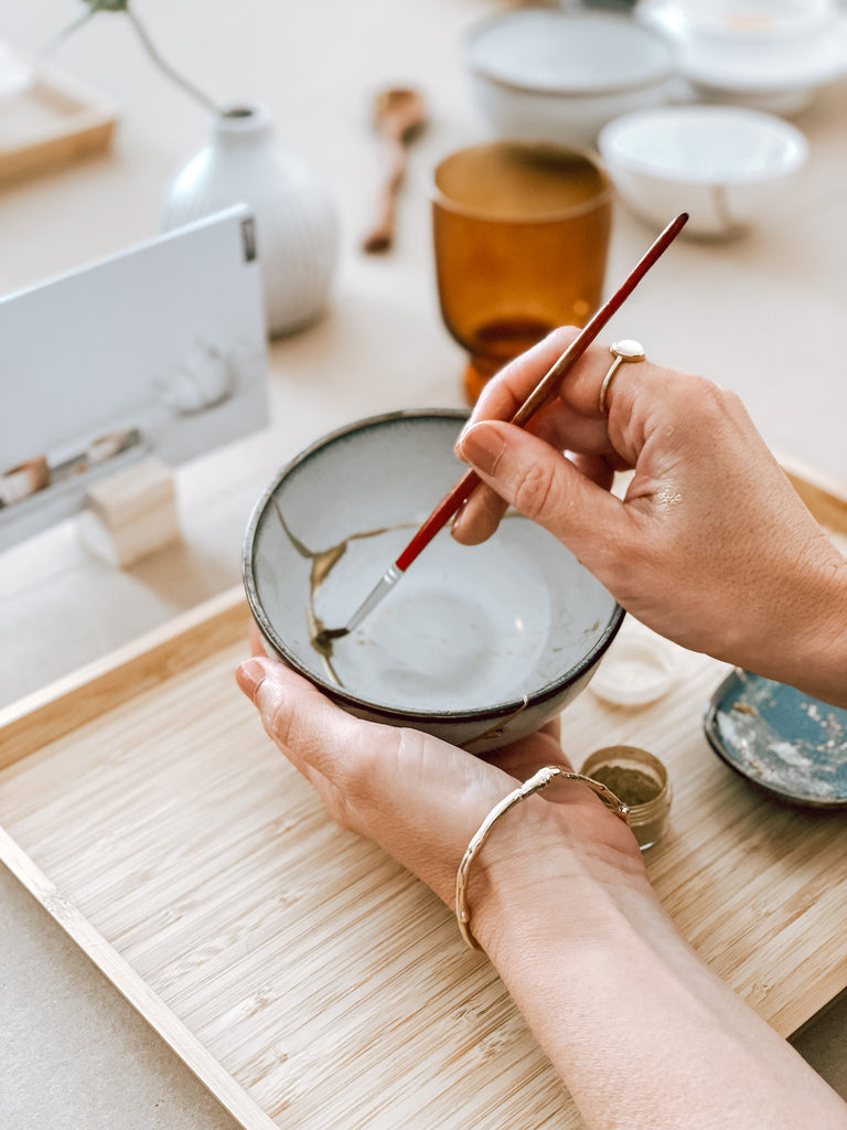 A woman's hands practising the art of Kintsugi on a small bowl. The woman is holding a paintbrush and adding gold to the cracks in the bowl.