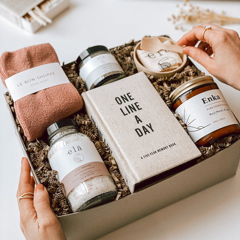 someone opening a byFoke Britta Gift Box which contains le Bon Shoppe socks, a One Line a Day Journal, a jar of Ela Life bath salts and Clay Mask set and an essential oil candle