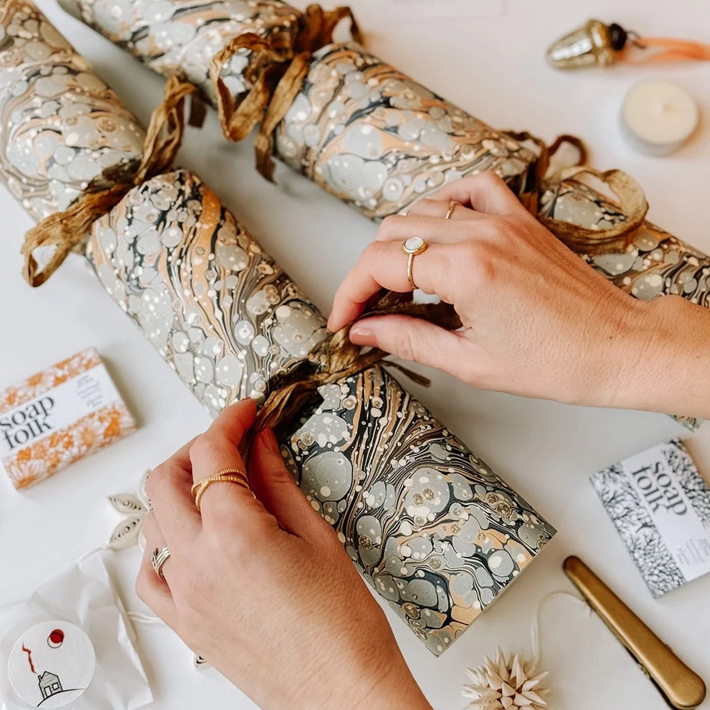 Two Marbled Fill your own Christmas Crackers. A pair of hands are tying the bow on one of the crackers, they are surrounded by little gifts which can go inside the crackers.