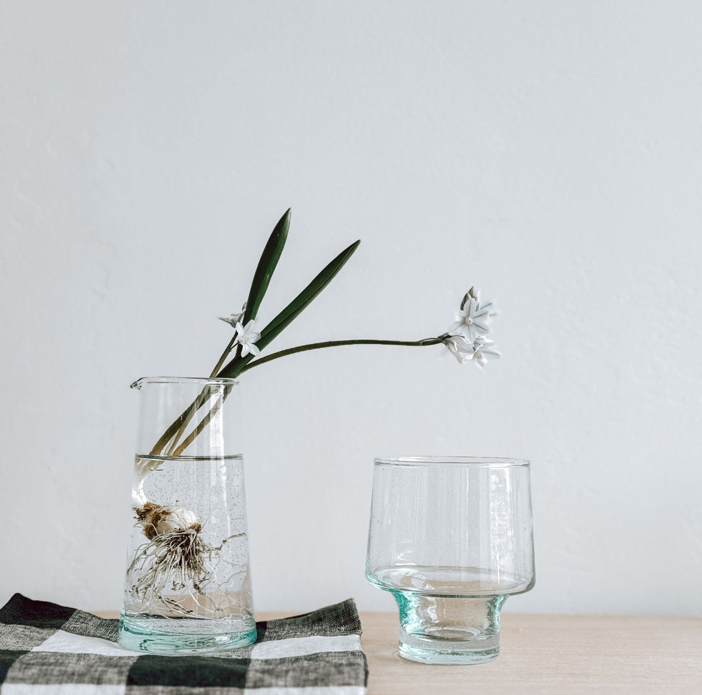 A short-stemmed low wine glass is on a wooden surface next to a taller, clear glass jug. The vase contains water, a plant bulb, and a white flower with green leaves, set on a checkered cloth.