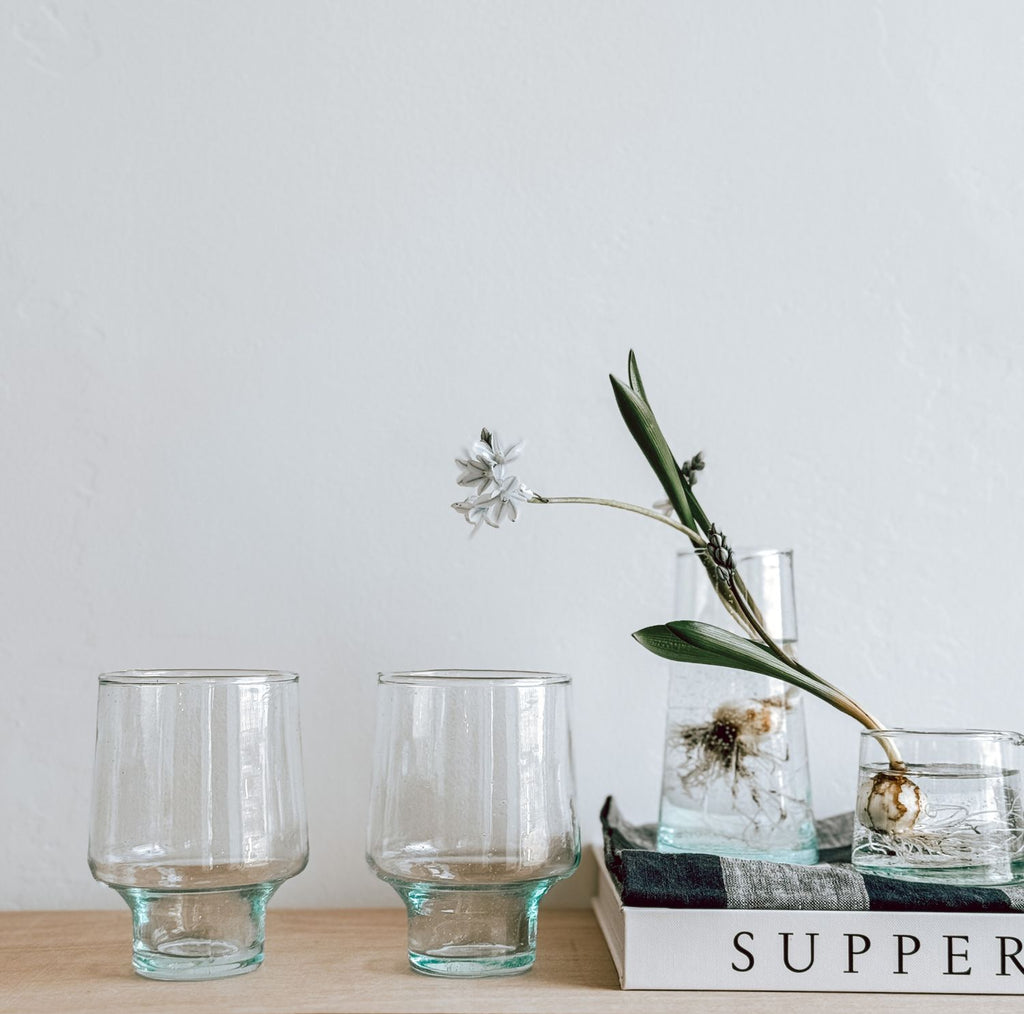 Two short-stemmed tall wine glasses on a wooden surface. To the right, there is a book topped with a gingham napkin with a glass jug on top containing water, a plant bulb, and a white flower with green leaves.