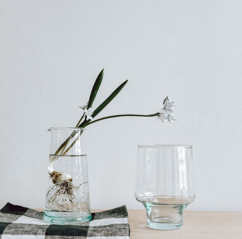 A short-stemmed tall wine glass is on a wooden surface next to a clear glass jug sitting on a folded gingham napkin. The vase contains water, a plant bulb, and a white flower with green leaves, set on a checkered cloth.