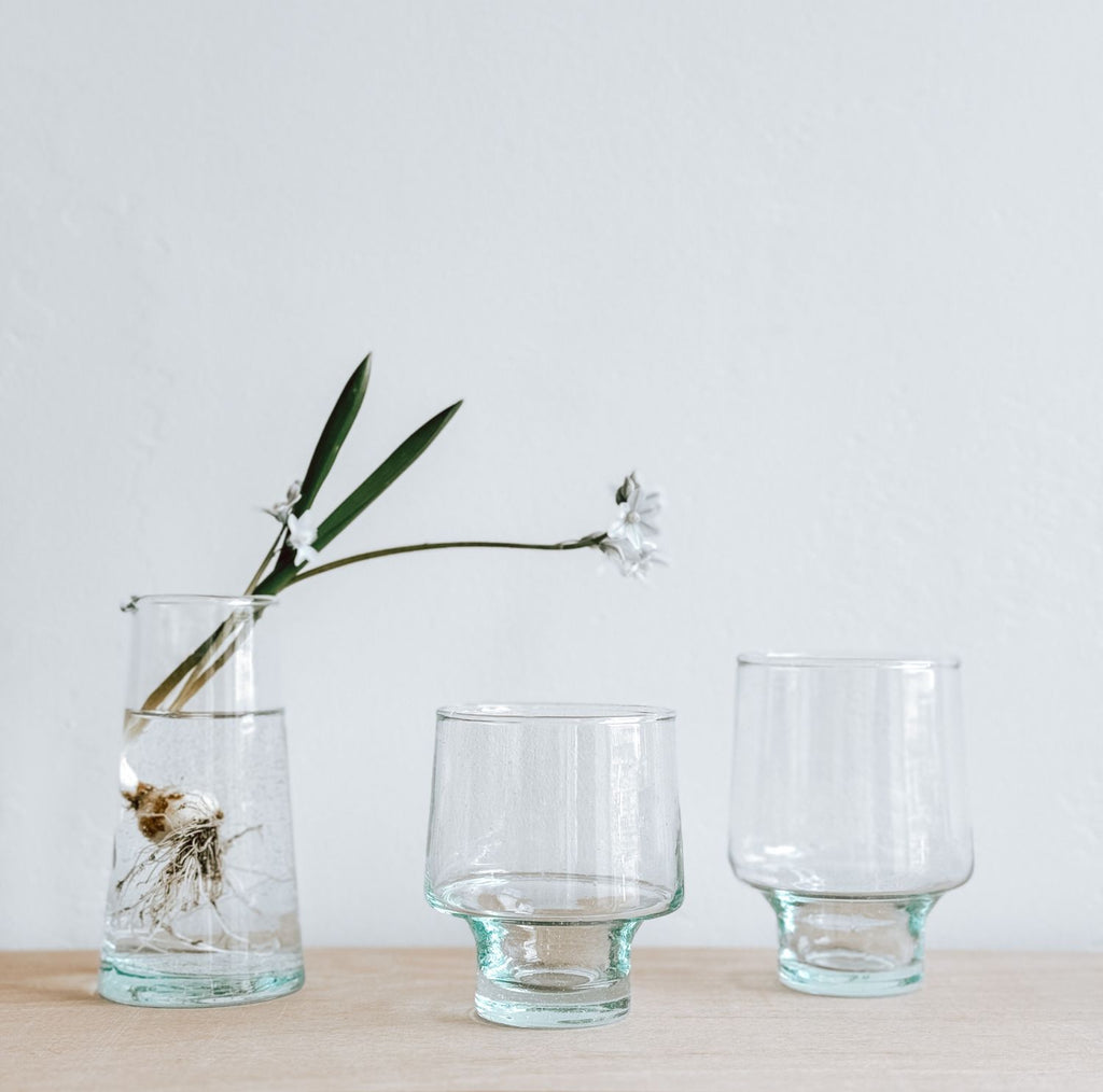 A pair of short-stemmed wine glasses one taller than the other on a wooden surface. Next to them, a tall, clear glass vase contains water, a plant bulb, and a white flower with green leaves.