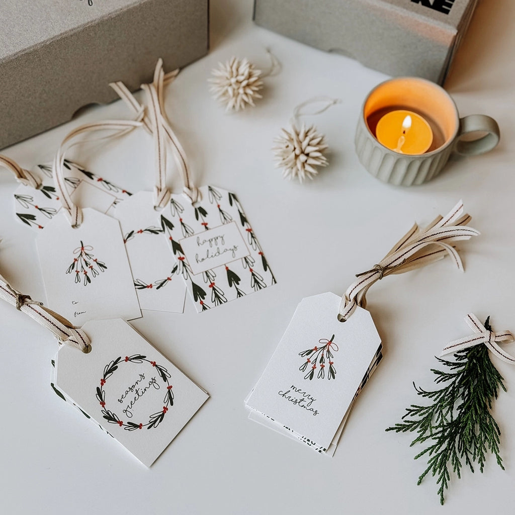 A set of byFoke Christmas Gift Tags with hand drawn illustrations are laying on a table with Christmas decorations, a sprig of pine and a lite tea light in the background.