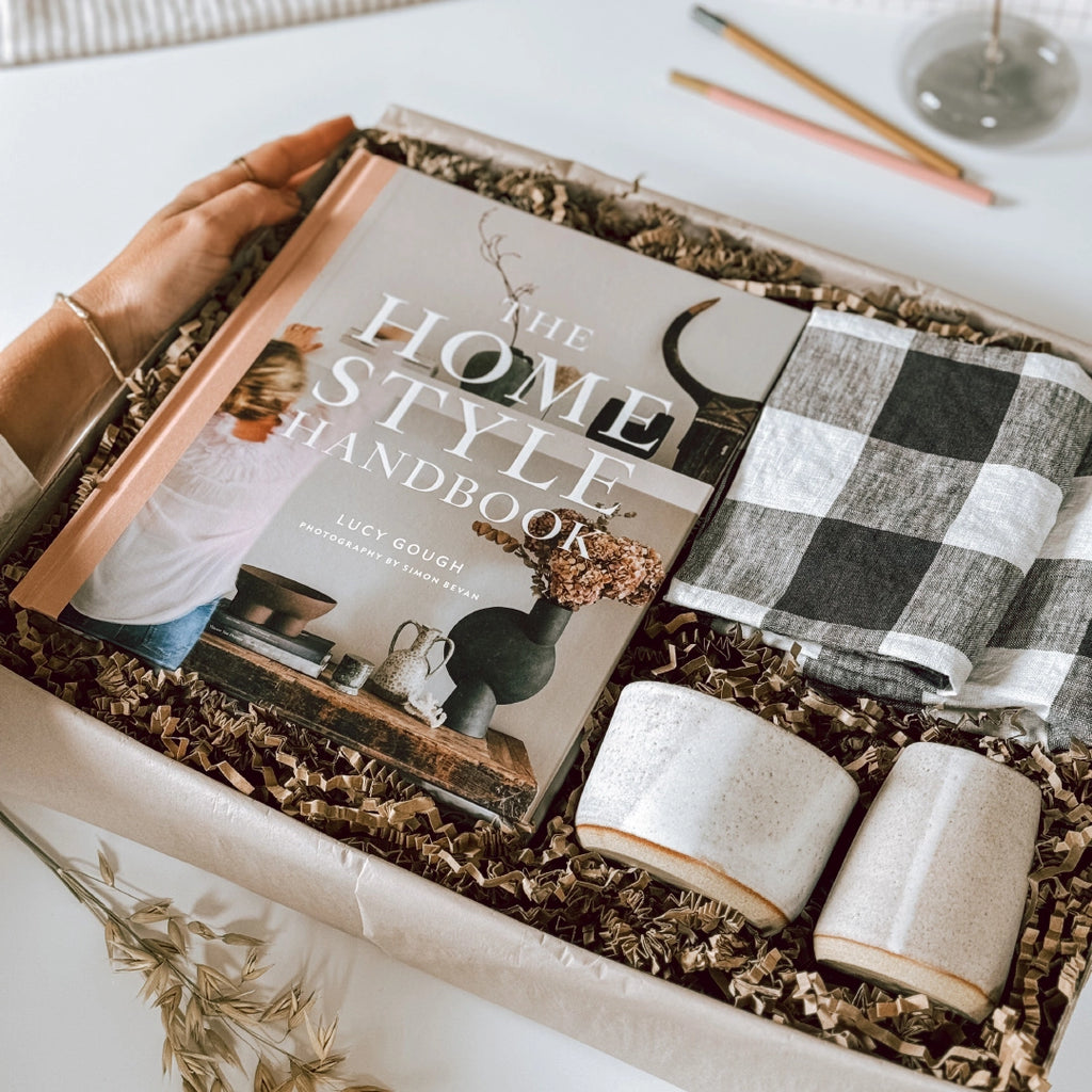 A byFoke gift box containing "The Home Style Handbook" by Lucy Gough, two folded black and white gingham napkins, a handmade ceramic jug and coffee bowl. A woman's hand is holding the edge of the box.