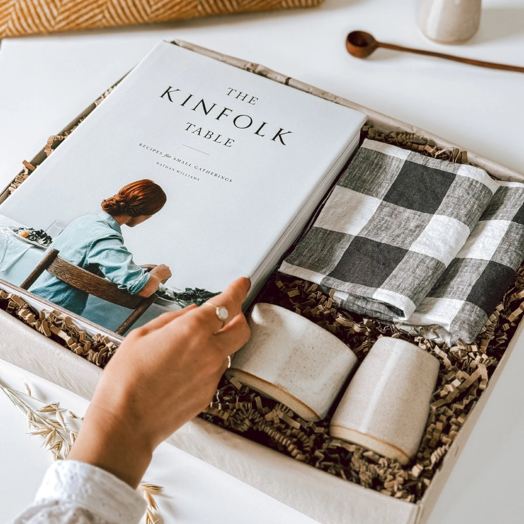 A byFoke gift box featuring "The Kinfolk Table" book by Nathan Williams, a set of black and white gingham napkins, and a small stoneware jug and bowl with an almond white glaze. A woman's hand is opening the book in the box.