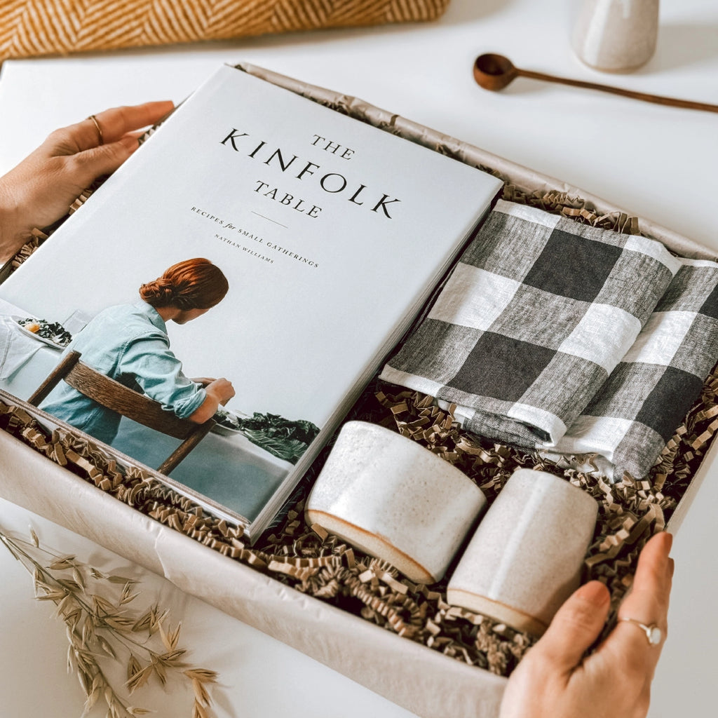 A byFoke gift box featuring "The Kinfolk Table" book by Nathan Williams, a set of black and white gingham napkins, and a small stoneware jug and bowl with an almond white glaze.  A woman's hands are holding the gift box.