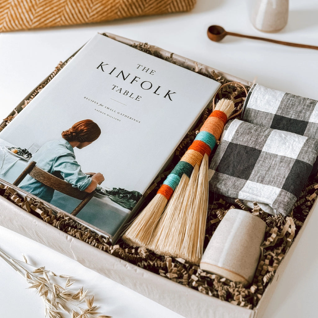 A byFoke gift box containing a copy of "The Kinfolk Table" book, two gingham linen napkins, a colourful tampico table broom, and a small ceramic jug. The gifts are beautifully arranged in the box, showcasing a cosy and rustic theme.