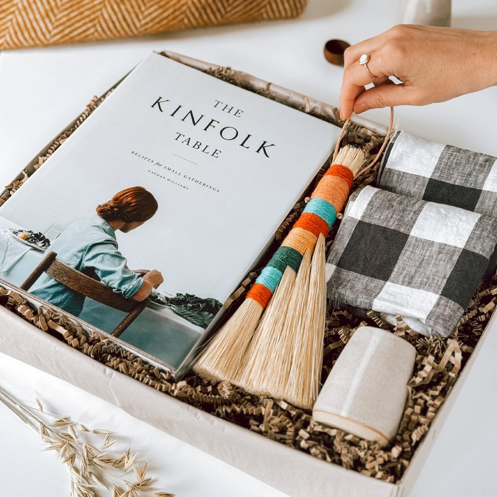 A byFoke gift box containing a copy of "The Kinfolk Table" book, two gingham linen napkins, a colourful tampico table broom, and a small ceramic jug. A woman's hand is lifting the tampico broom out of the box.