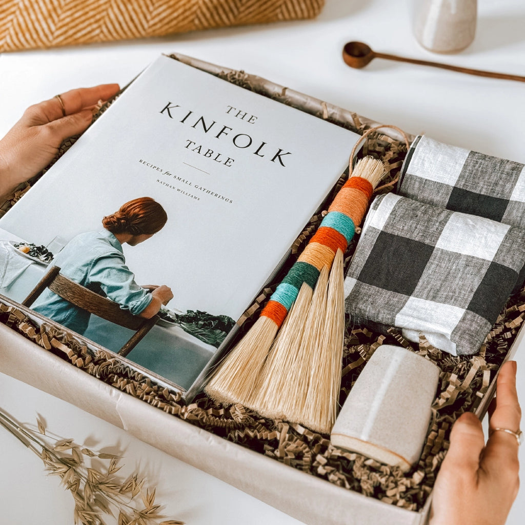 A byFoke gift box containing a copy of "The Kinfolk Table" book, two gingham linen napkins, a colourful tampico table broom, and a small ceramic jug. A woman's hands are holding the box.