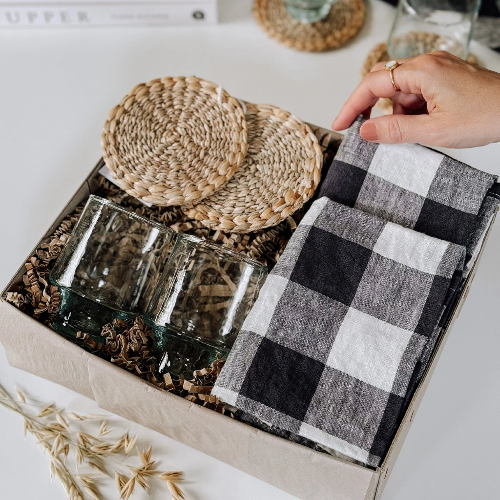 byFoke Greta gift box containing woven coasters, two wine glasses, and two folded black-and-white gingham napkins, all nestled in natural packing material. A hand is lifting the corner of one of the napkins.