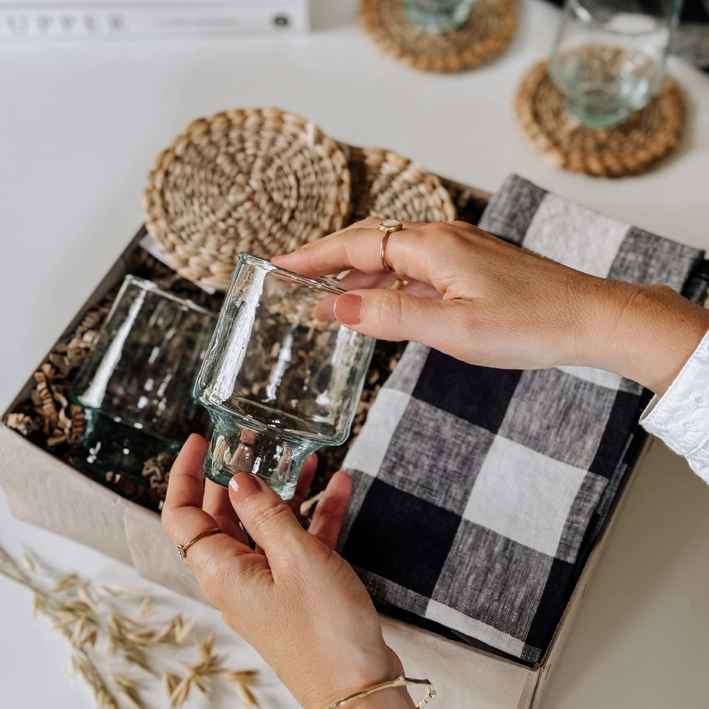 byFoke Greta gift box containing woven coasters, two wine glasses, and two folded black-and-white gingham napkins, all nestled in natural packing material. Hands are lifting one of the hand-blown glasses out of the gift box.