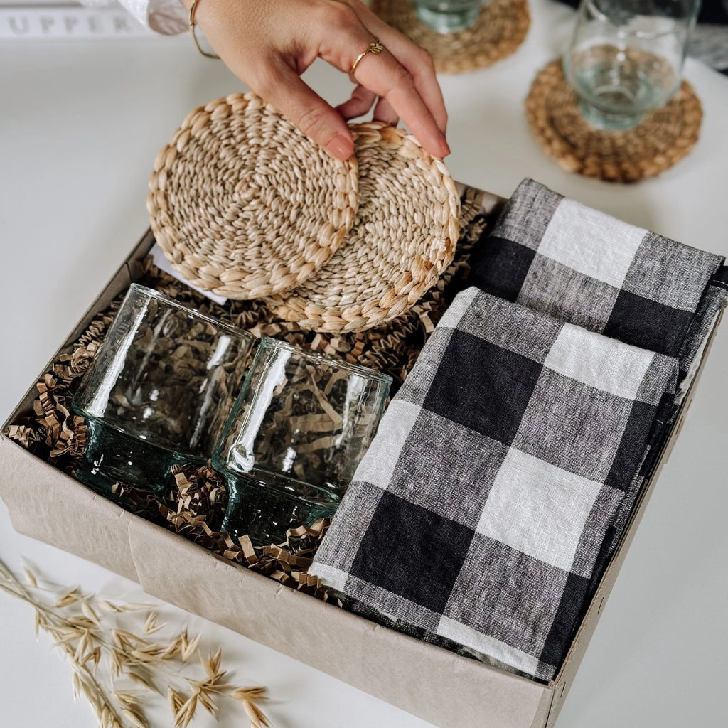 byFoke Greta gift box containing woven coasters, two wine glasses, and two folded black-and-white gingham napkins, all nestled in natural packing material. A hand is lifting the coasters out of the gift box.