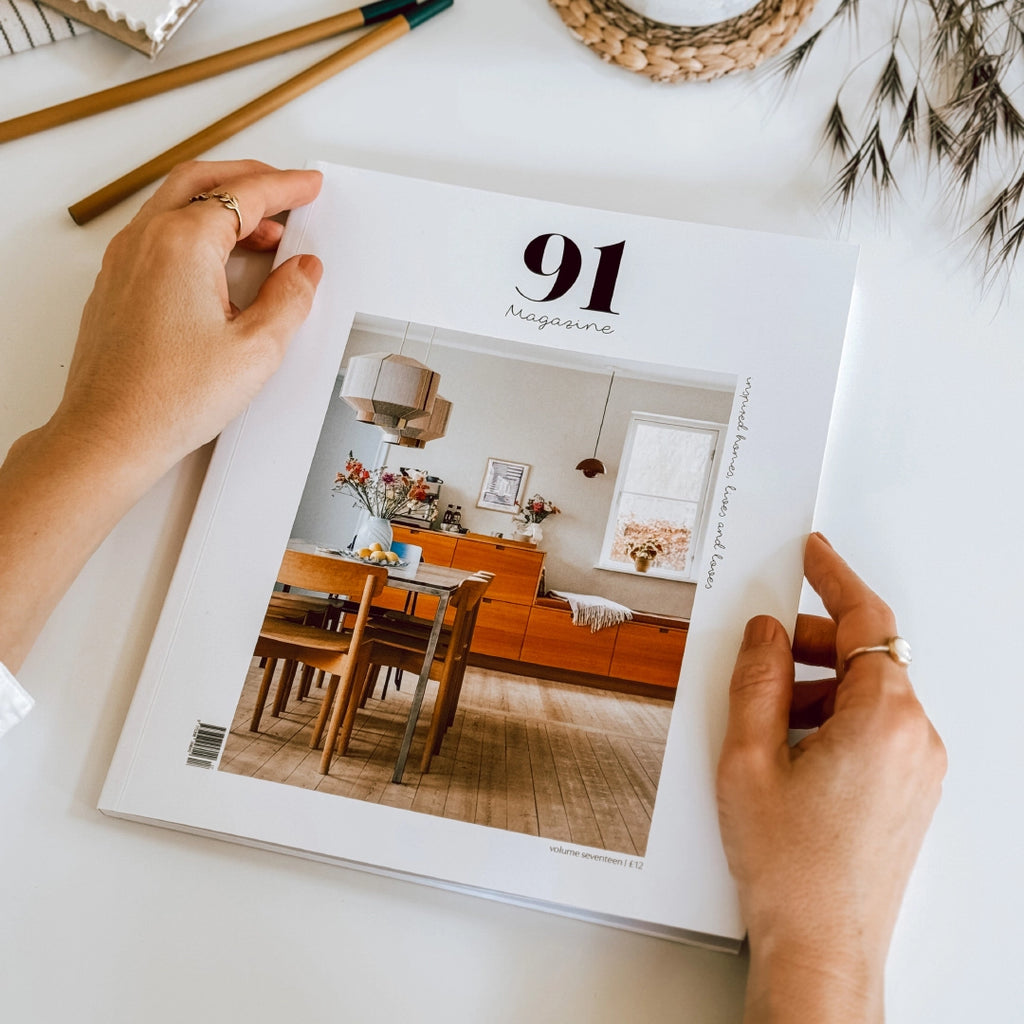 Hands holding 91 Magazine Volume 17 on a table with pencils and dried grasses in the background, byFoke