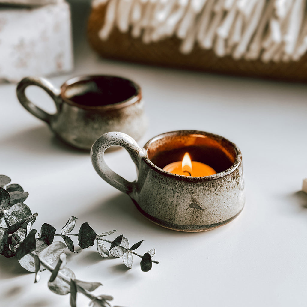 A beautiful pair of stoneware candle holders, with a little handle and a speckled tawny glaze. There is a lit tea light glowing in one of the holders.