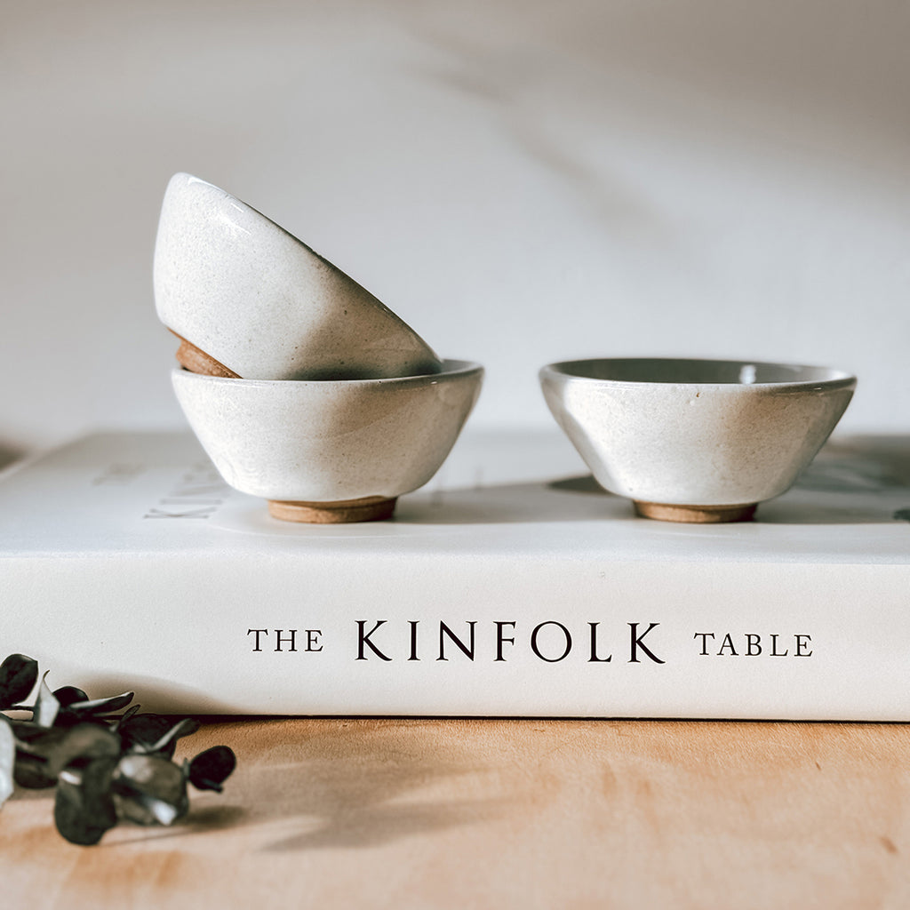 A set of three small stoneware footed bowls with a cream glaze. The bowls are sitting on top of The Kinfolk Table book.