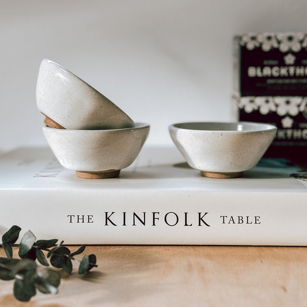 A set of three small stoneware footed bowls with a cream glaze. The bowls are sitting on top of The Kinfolk Table book, with some Blackthorn Salt in the background.