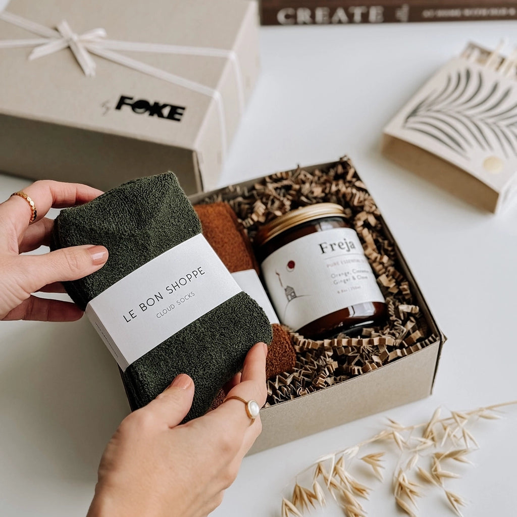 The Chloe Gift Box byFoke, laying open with the lid to one side. A woman's hands are holding up a pair of forest Cloud socks which are in the gift with an essential oil candle.