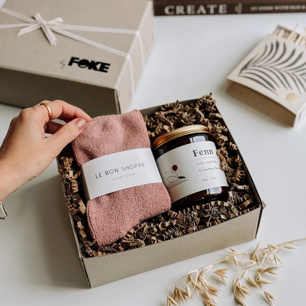 byFoke's Chloe Gift Box, opened with the lid to one side. A woman's hand is holding up the corner of the mulberry Cloud socks which are sitting next to a Fenn essential oil candle inside the gift box.
