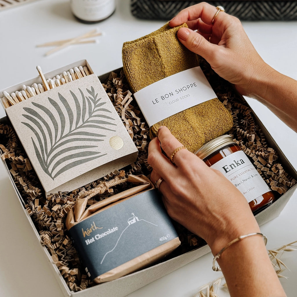 e Bon Shoppe socks, an Essential Oil Soy Wax Candle, a box of luxury matches in a letterpress printed box, and Hearth Hot chocolate are all neatly arranged in a gift box on the table