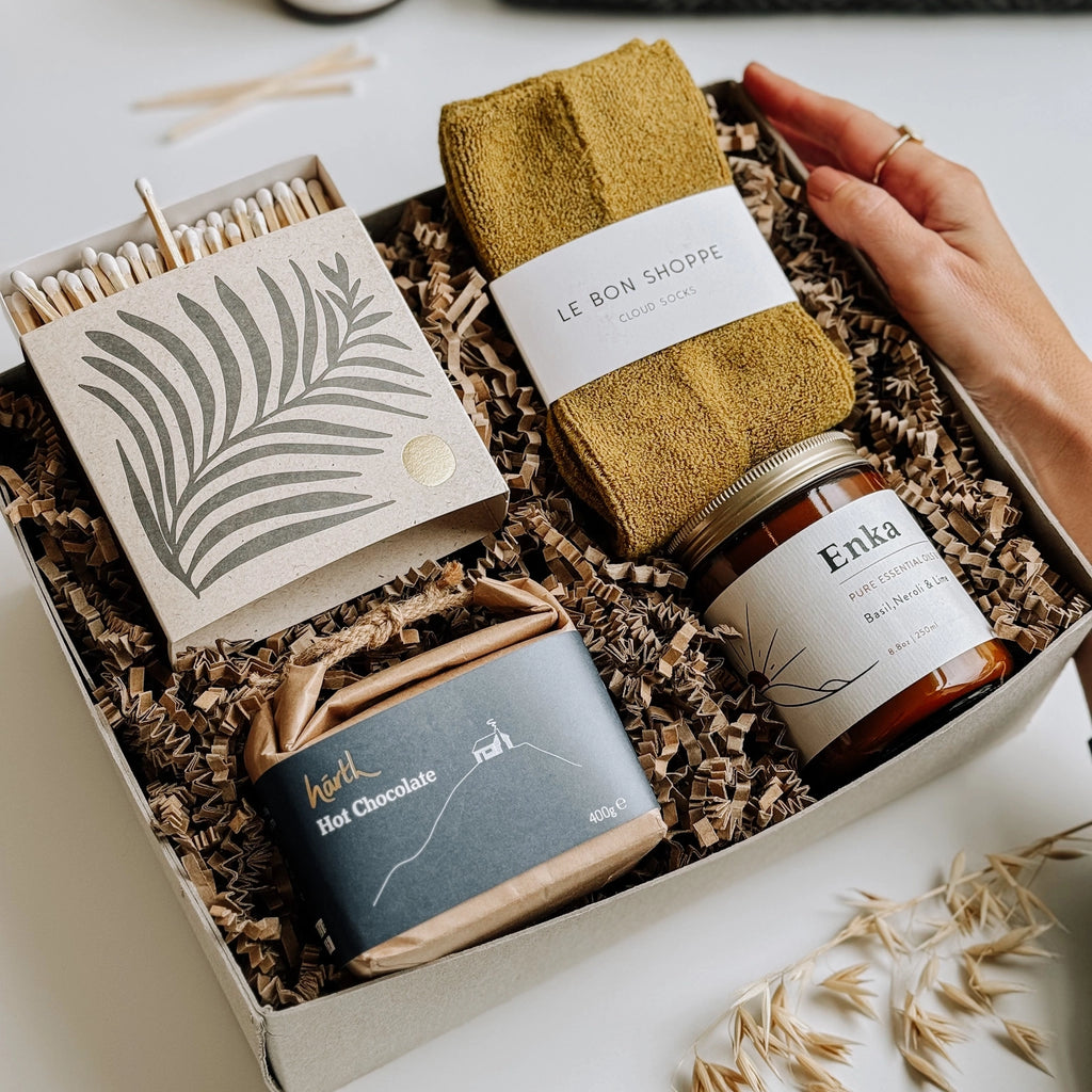 On the table, there's a gift box with Le Bon Shoppe socks, an Essential Oil Soy Wax Candle, a box of luxury matches in a letterpress printed box, and some Hearth Hot chocolate.