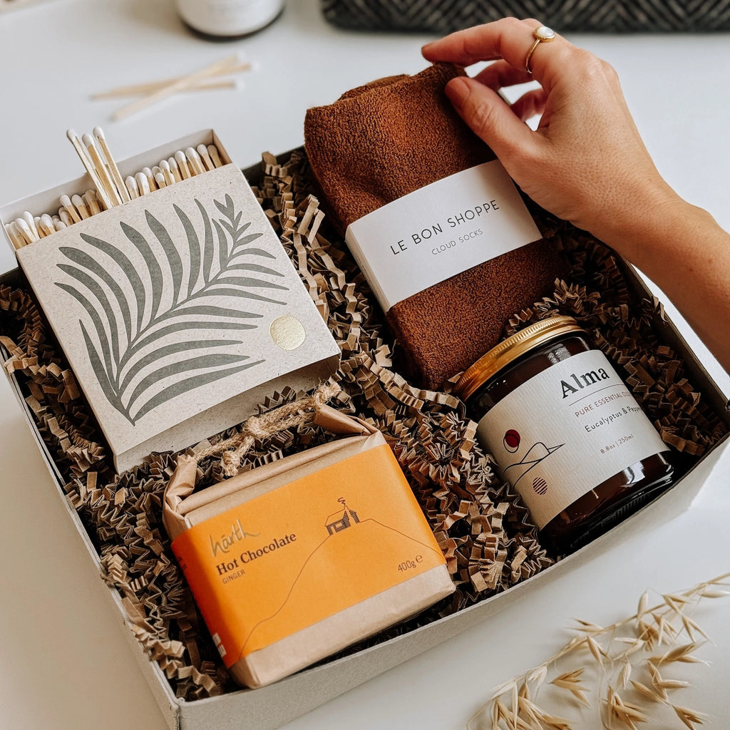 You'll find a gift box on the table, complete with Le Bon Shoppe socks, an Essential Oil Soy Wax Candle, a box of luxury matches in a letterpress printed box, and Hearth Hot chocolate.