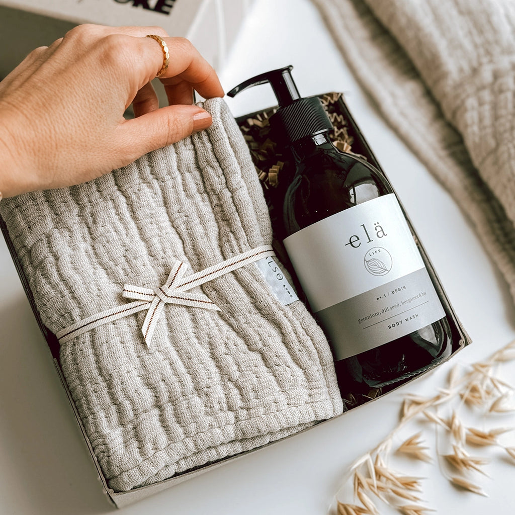 A woman's hand is lofting the corner of a stone coloured wash cloth, which is sitting next to a glass bottle of Ela Life Body Wash inside an Una Gift Box.