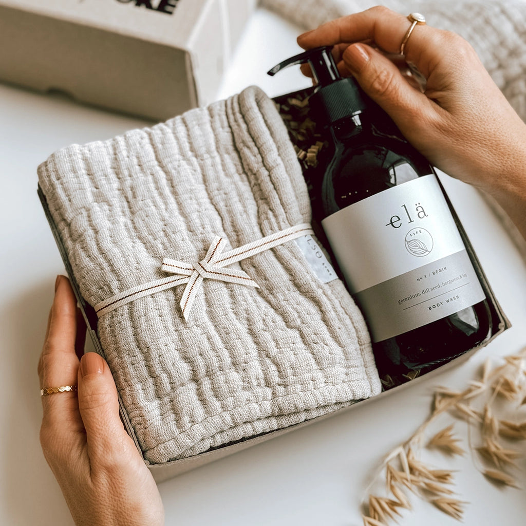 A woman's hands are holding the Una Gift Box, which is open , inside is a glass bottle of Ela Life Body Wash and a soft stone coloured wash cloth.