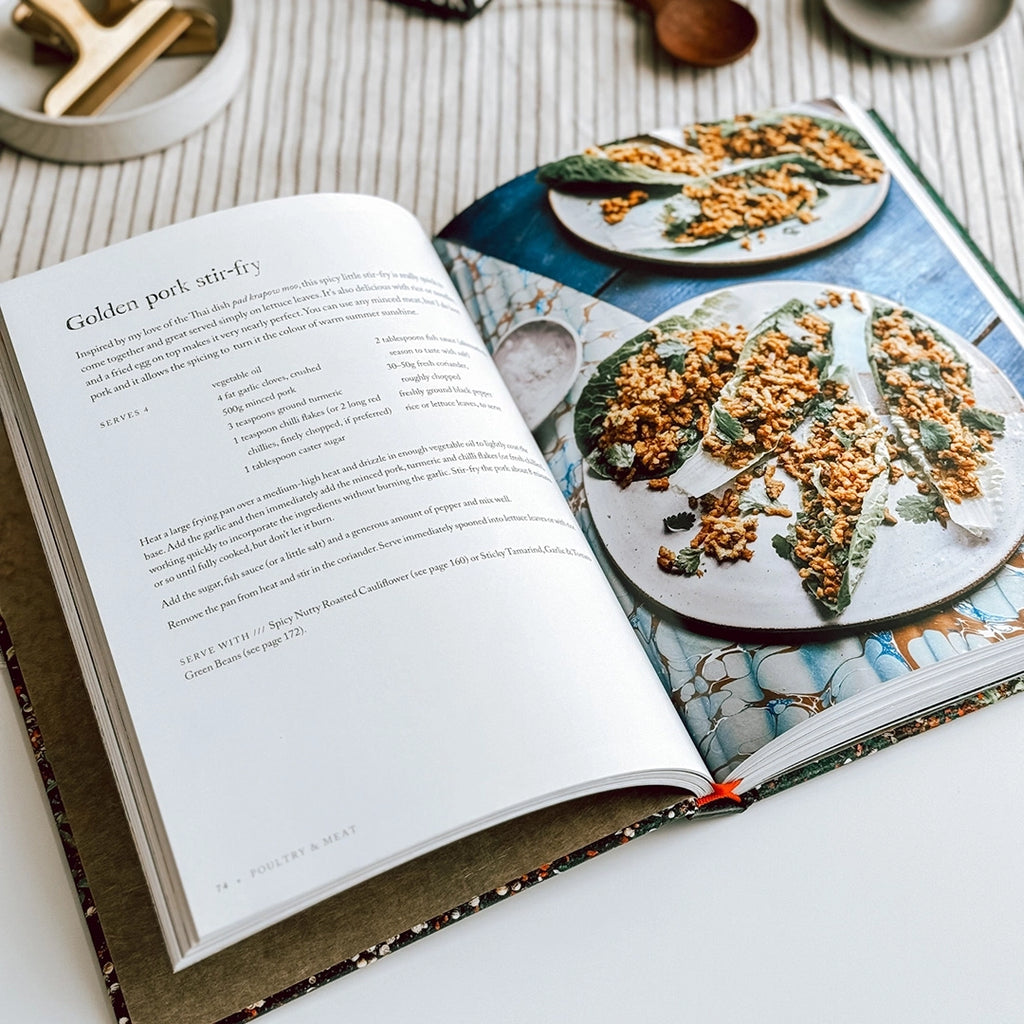 The table hosts an open copy of Sabrina Ghayour's cookbook "Persiana Everyday" with the Golden pork stir-fry recipe in the photo.