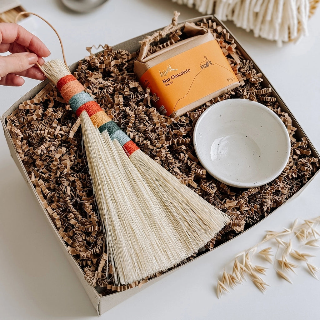 A woman's hand is lifting the top of a woven Tampico brush with a striped wool handle, which is inside a byFoke Hanne Gift Box which also contains some Harth Hot Chocolate and a small ceramic bowl.and a small ceramic bowl.