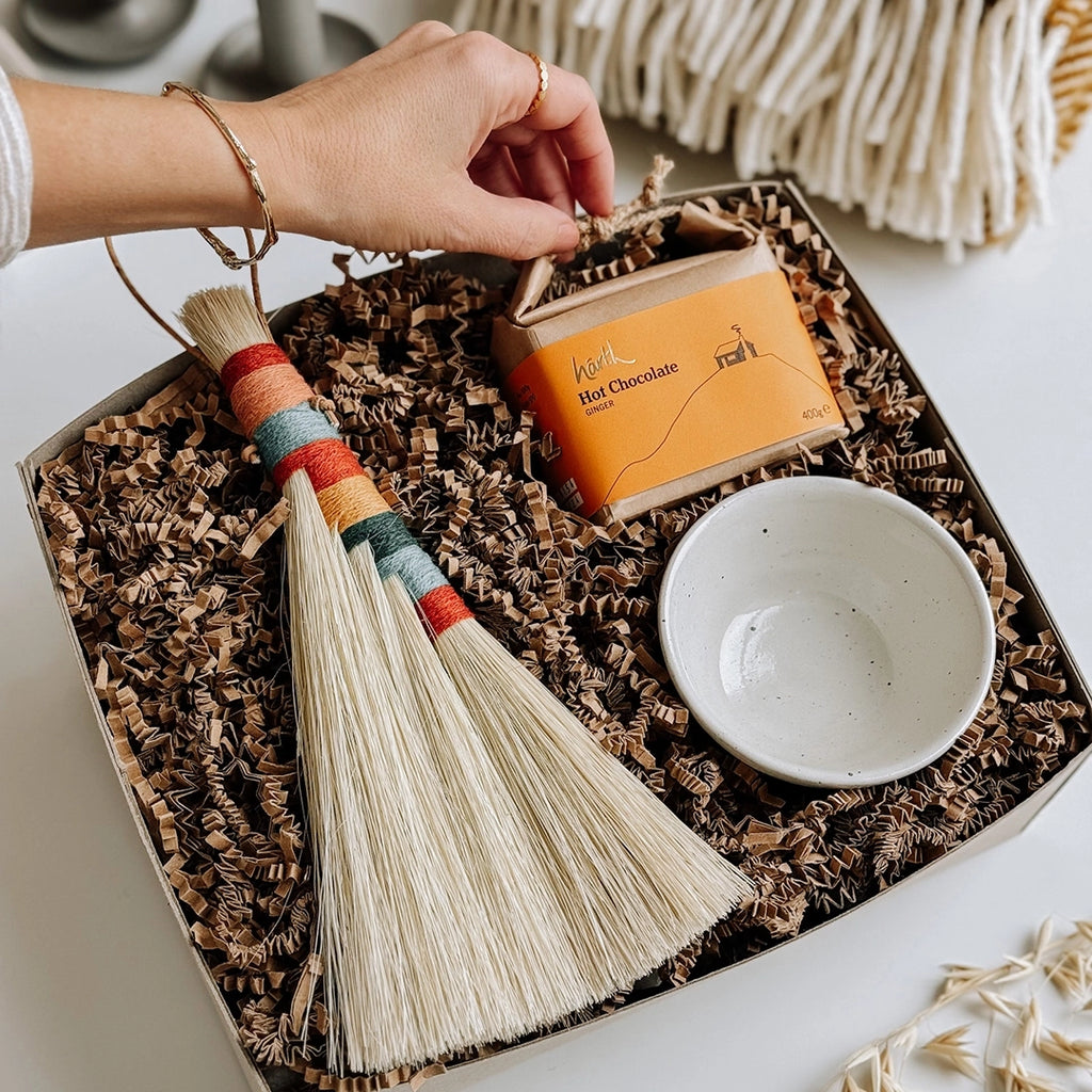 A woman's hand is lifting the corner of the Harth Hot Chocolate out of byFoke's Hanne Gift Box, which also contains a woven Tampico brush with a striped wool handle and a small ceramic bowl.