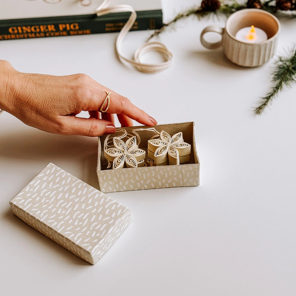 A hand is opening a painted paper box which is filled with a set of 6 cream quilled paper star decorations. Pine cones and pine sprigs are in the background.