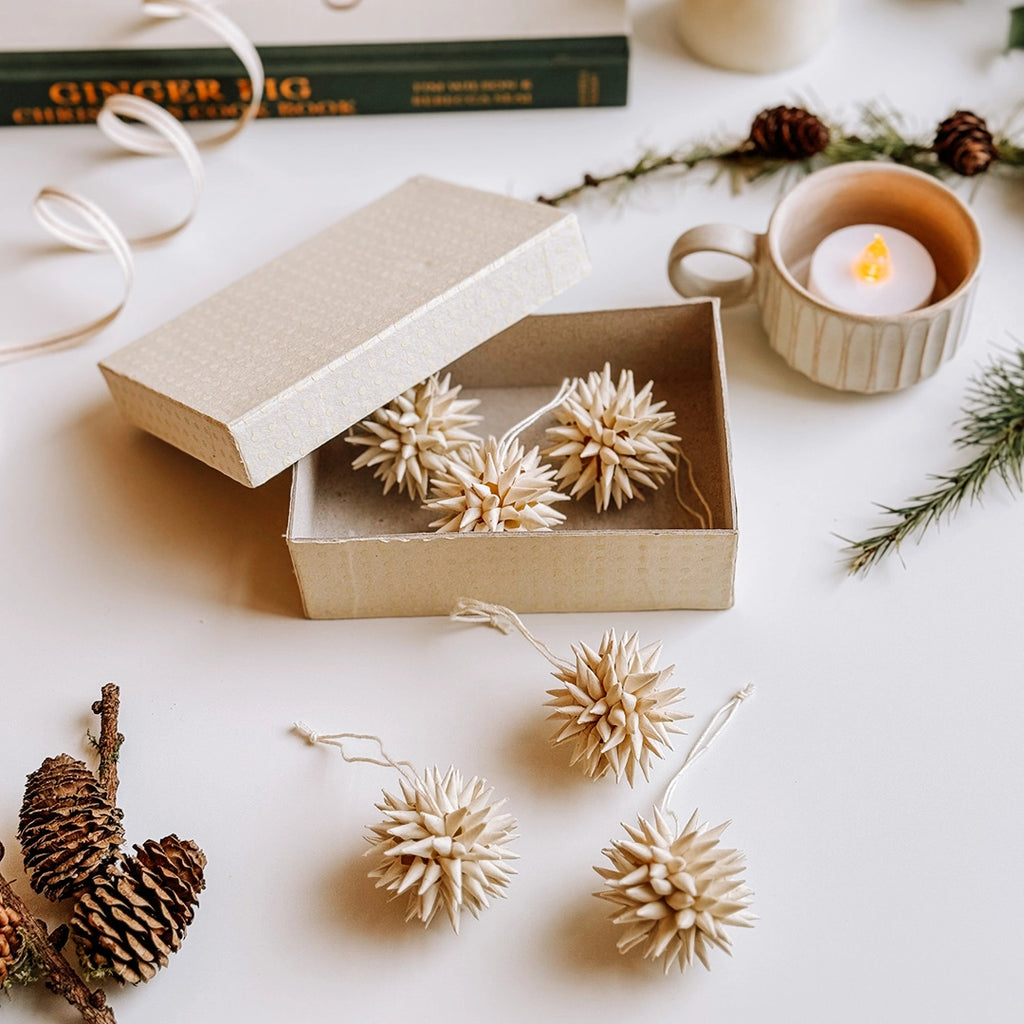 A box of cream quilled paper Christmas decorations. a tea light, pine cones and a Christmas recipe book are in the background.